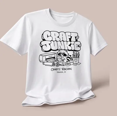 Cartoon raccoon graphic with text 'CRAFT JUNKIE CRAFTY RACOON PAHOA, HI' in black and white — printed on a unisex t-shirt (angled view)